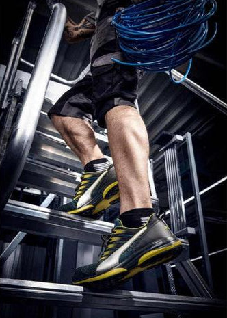 A person wearing black shorts and a pair of Puma ELEVATE KNIT safety shoes with a blue and gray color scheme, is depicted climbing a metal staircase with a blue wire and a black bag on his shoulder.